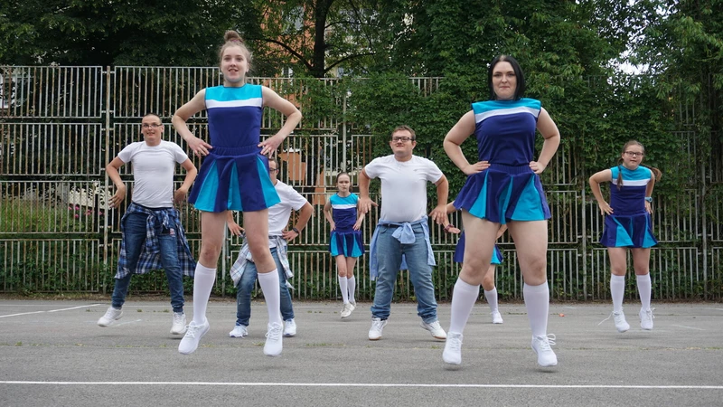 Kilkoro dziewcząt i chłopców w niebieskich strojach tanecznych w stylu czirliderek tańczy na szkolnym boisku sportowym.