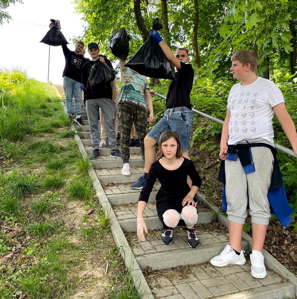 Młodzież trzyma w dłoniach worki czarne, foliowe. Stoją w otoczeniu drzew, prawdopodobnie w parku.