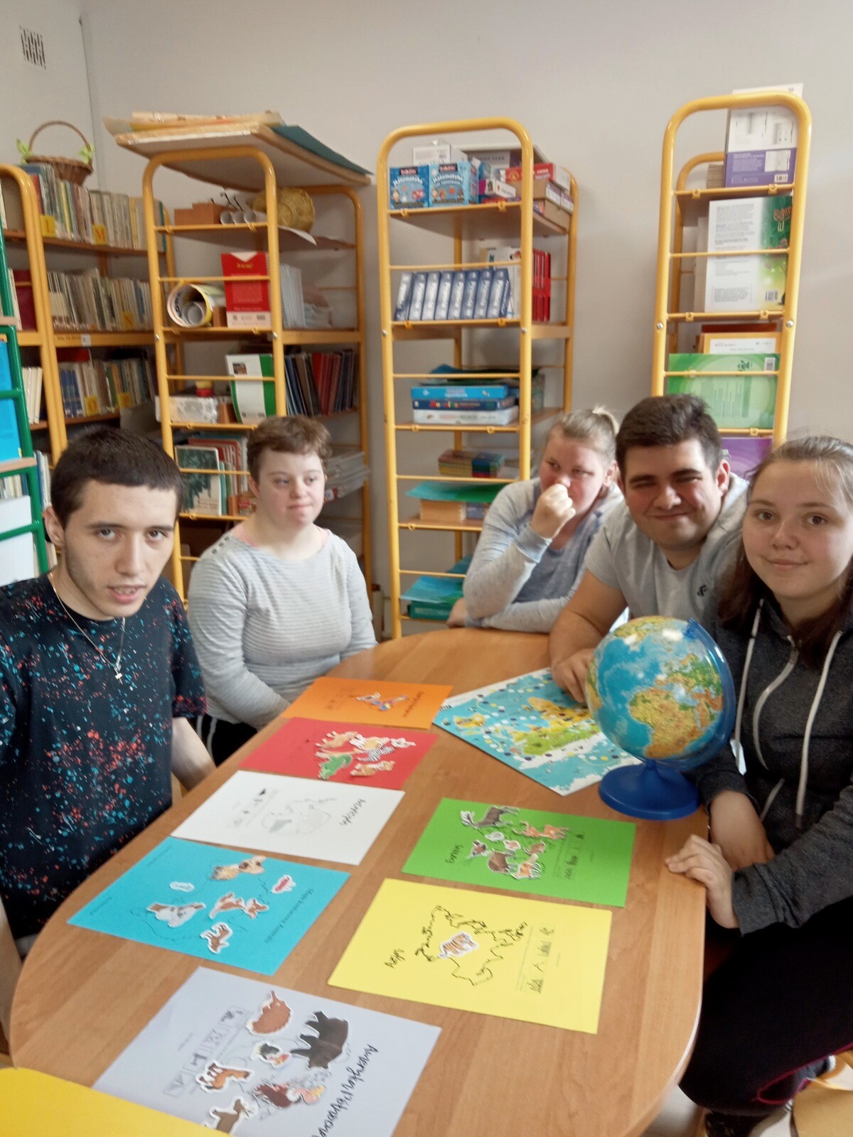 Grupa uczniów siedzi przy stoliku w bibliotece szkolnej, W tle widac stojące regały z książkami.