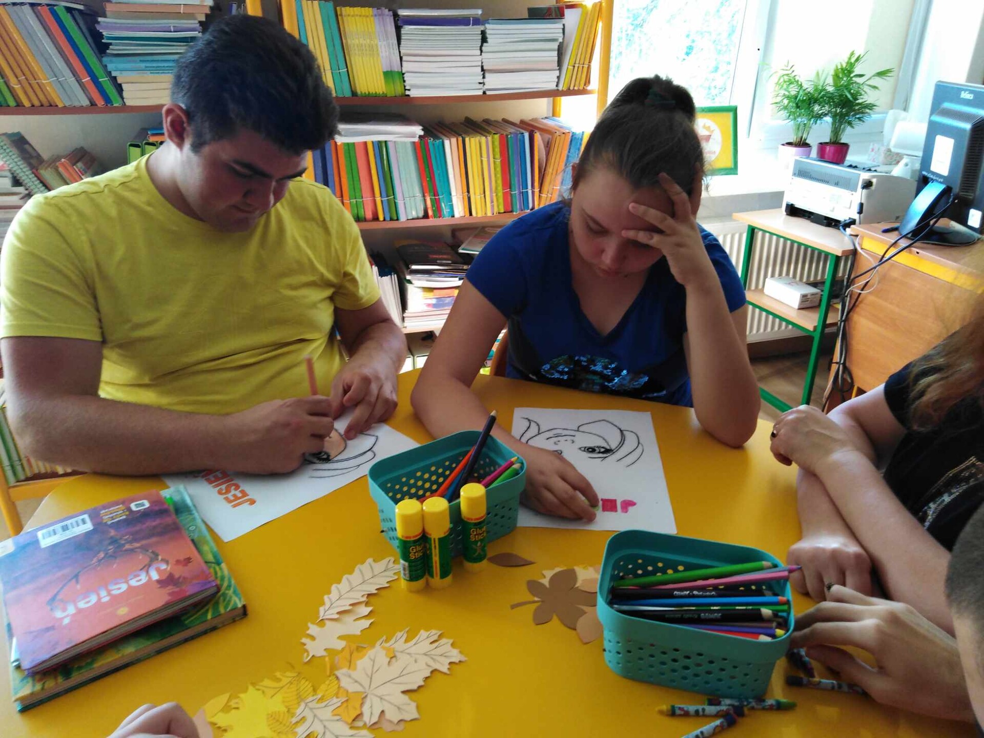 Uczniowie siedzą  przy stoliku w bibliotece szkolnej. W tle widać regały z książkami.