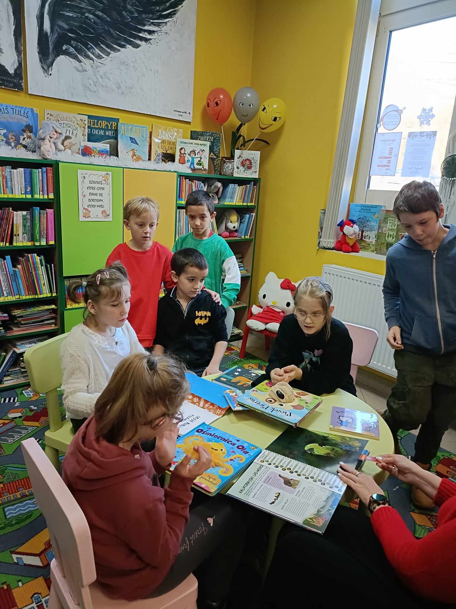 Grupa uczniów siedzi przy stoliku w bibliotece, przed nimi rozłożone sa na stole rózne ksiązki. Wokół stolika stoją też dzieci i patrzą na ksiązki.