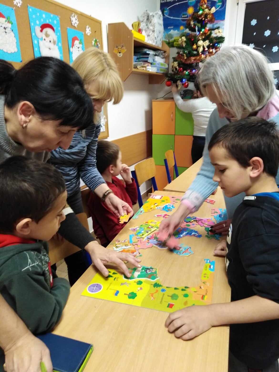 Grupa dzieci z dwiema nauczycielkami pochylają się nad stołem u układają puzzle.