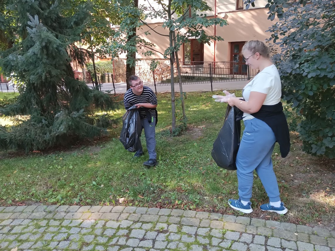 Dziewczyna i chłopiec trzymają w dłoniach czarne worki foliowe, są w ogrodzie, zbierają śmieci.