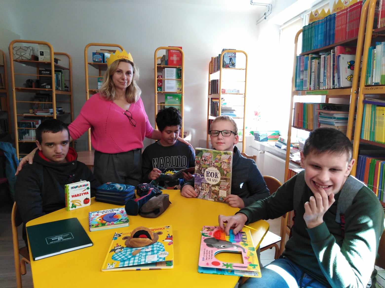 Uczniowie siedzą przy żółtym stoliku w bibliotece szkolnej, na stoliku leżą ksiązki, które uczniowie przeglądają. Nauczycielka stoi obok stolika, na głowie ma koronę.