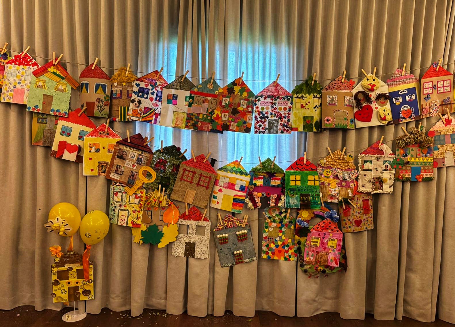 Kolorowe papierowe domki zawieszone na zasłonach - wystawa prac plastycznych.