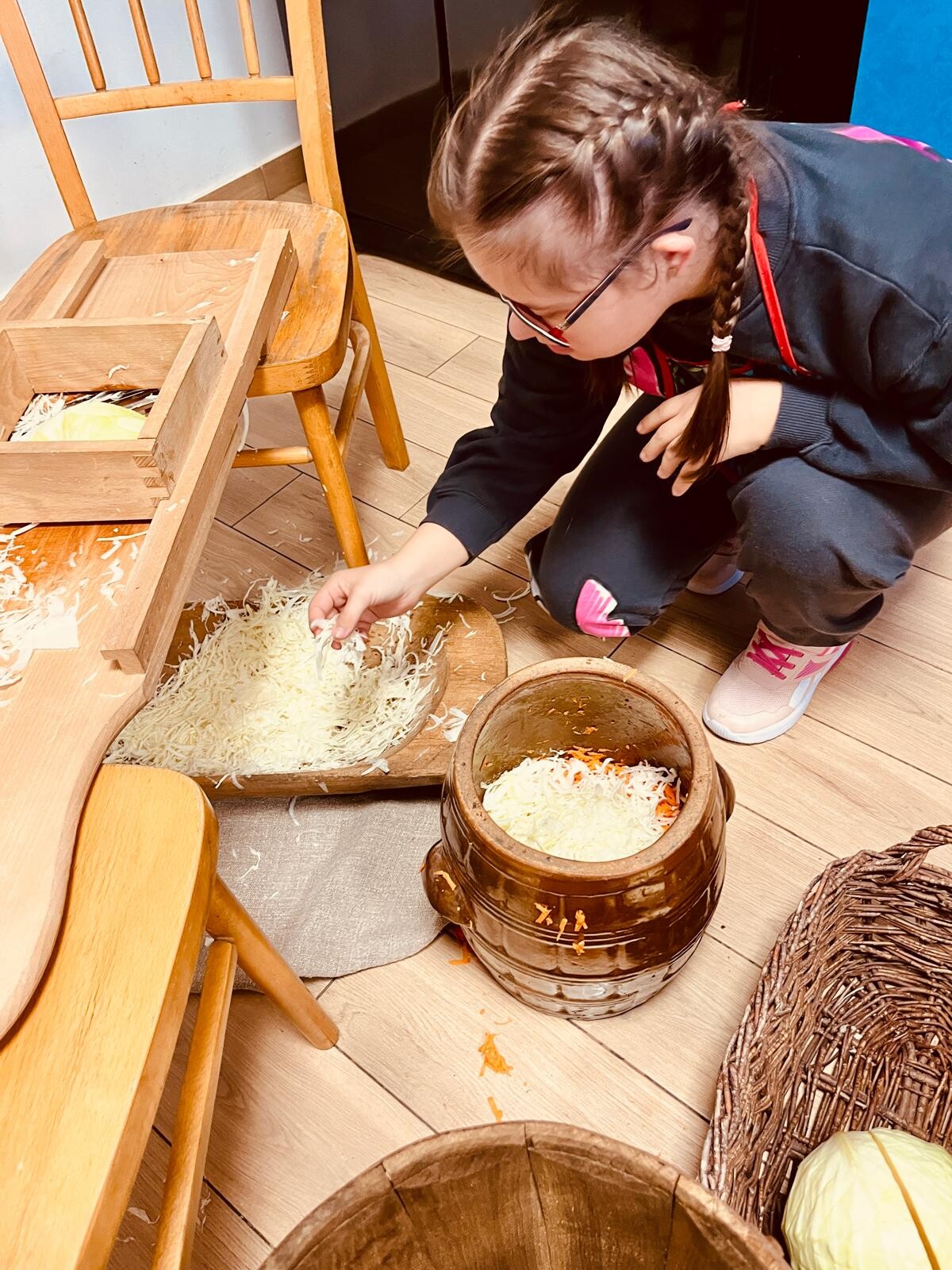 Dziewczynka nachyla się nad garnkiem wypełnionym poszatkowaną kapustą. Obok stoi szatkownica do kapusty.