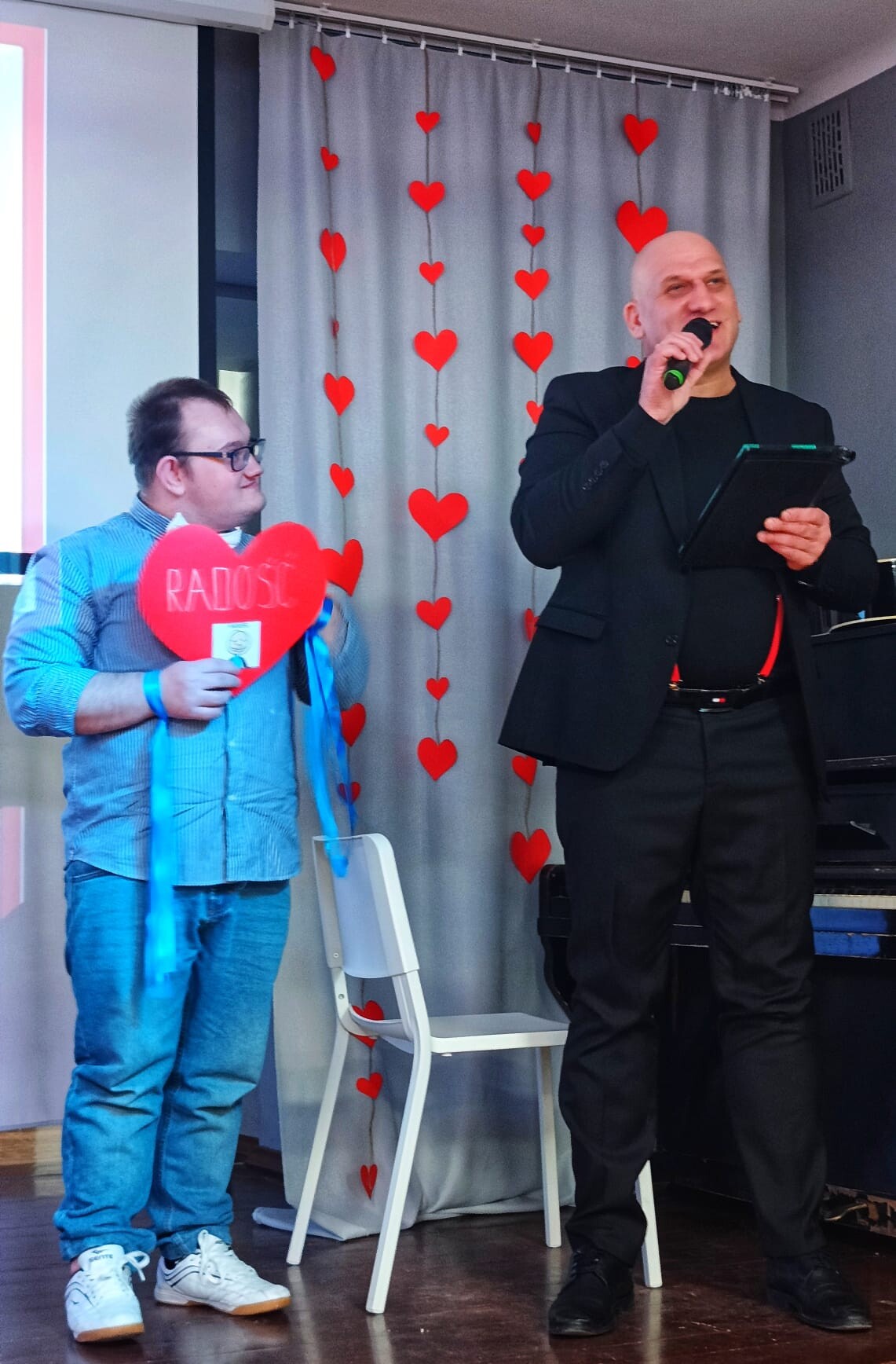 Uczeń czyta tekst z kartki w kształcie serca, obok stoi nauczyciel z mikrofonem w dłoni.