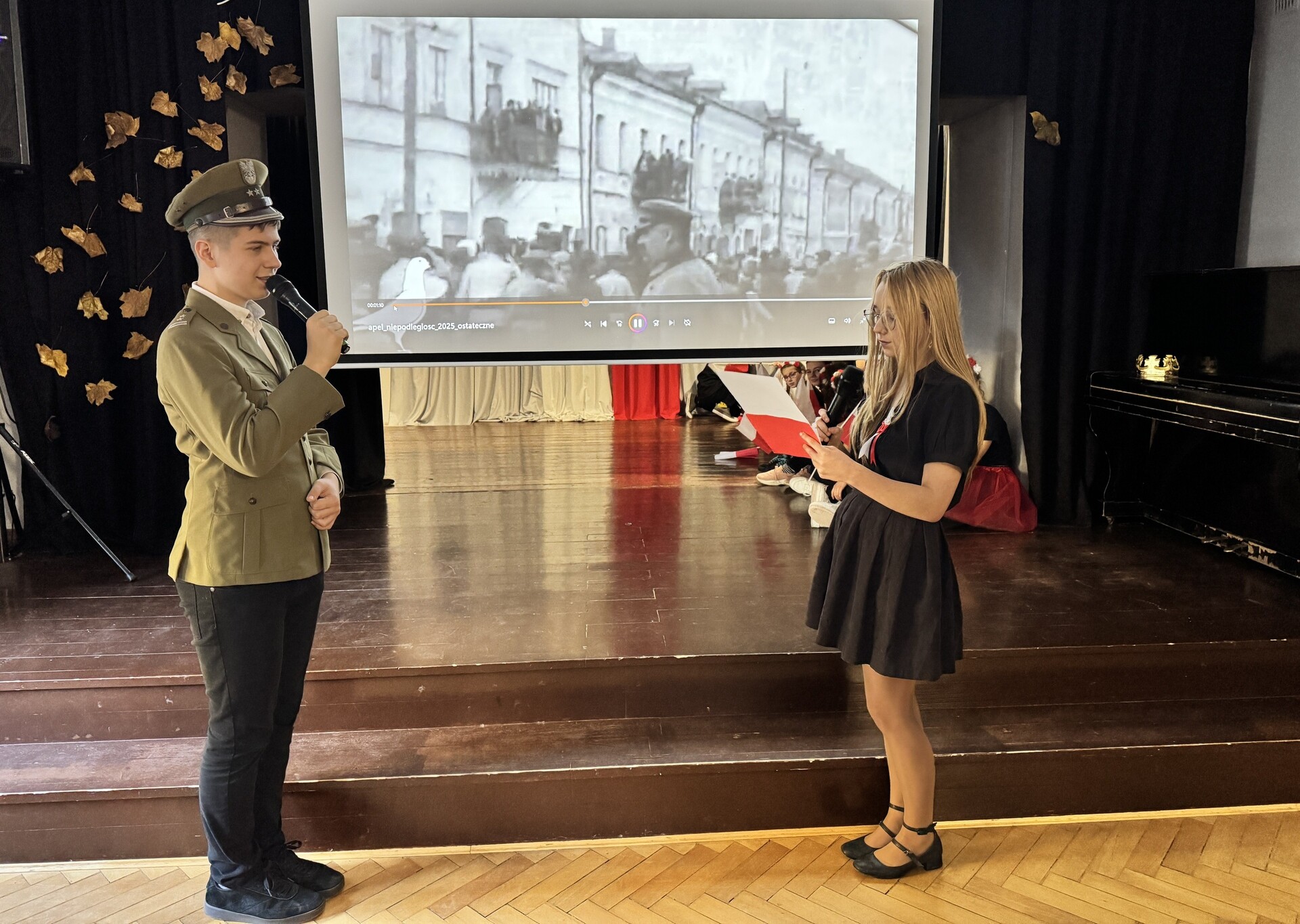 Uczeń ubrany w mundur mówi do mikrofonu, stoi na przeciw uczennicy ubranej w strój galowy. W tle wyświetlany jest slajd z Józefem Piłsudskim.