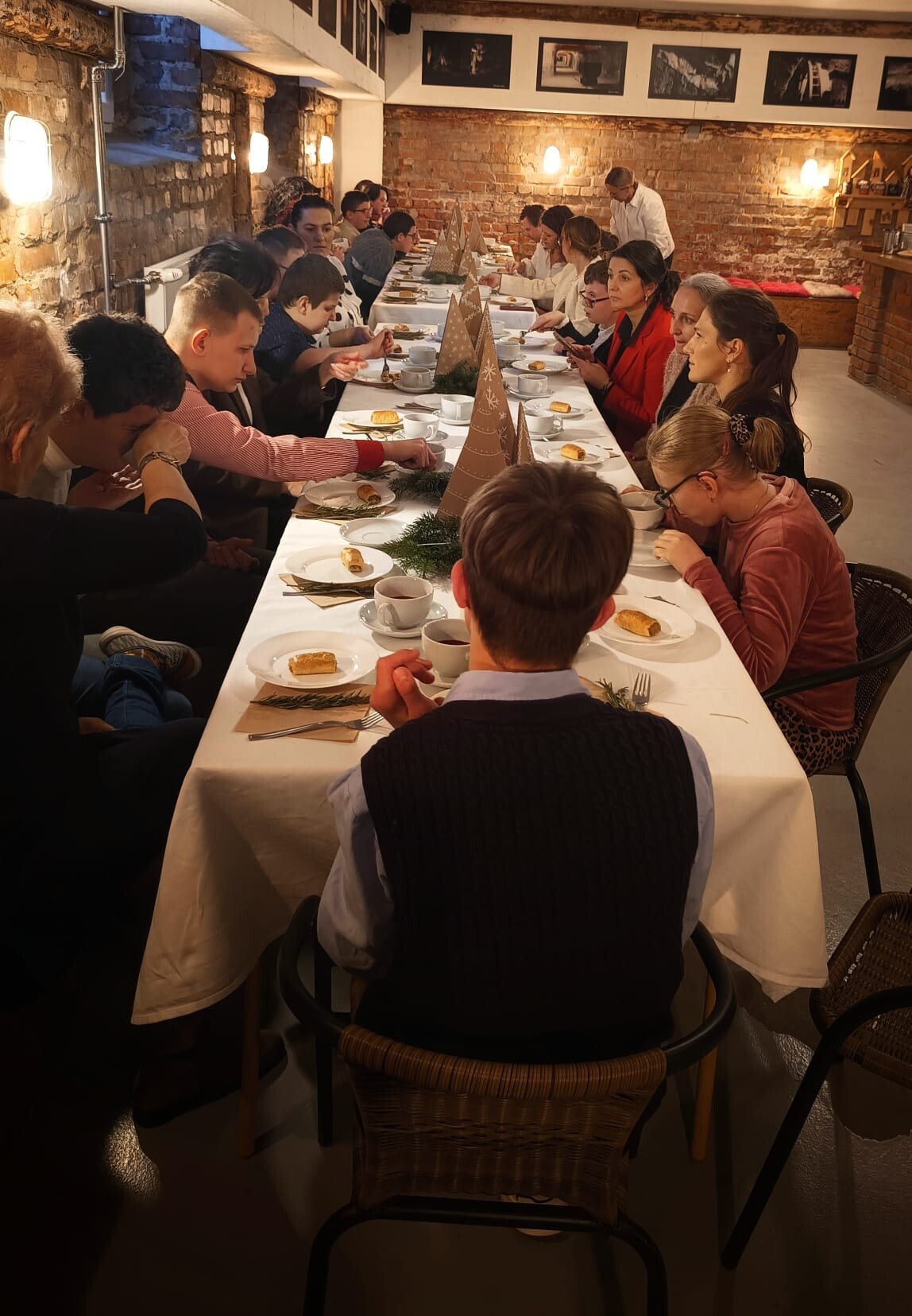 Uczniowie w odświetnych ubraniach siedzą wokół przystrojonego białym obrusem stołu wigilijnego.