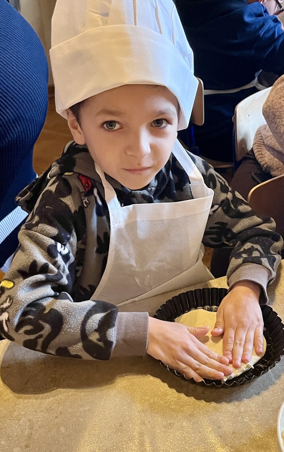 Chłopczyk w czapce kucharskiej i fartuchu siedzi przy stole, w dłoniach trzyma mała formę na ciasto.
