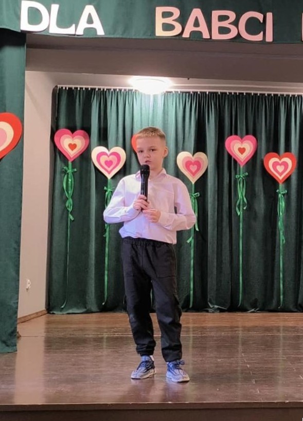 Mały chłopiec stoi na scenie, w  dłoni trzyma mikrofon. W tle widać serca z papieru.