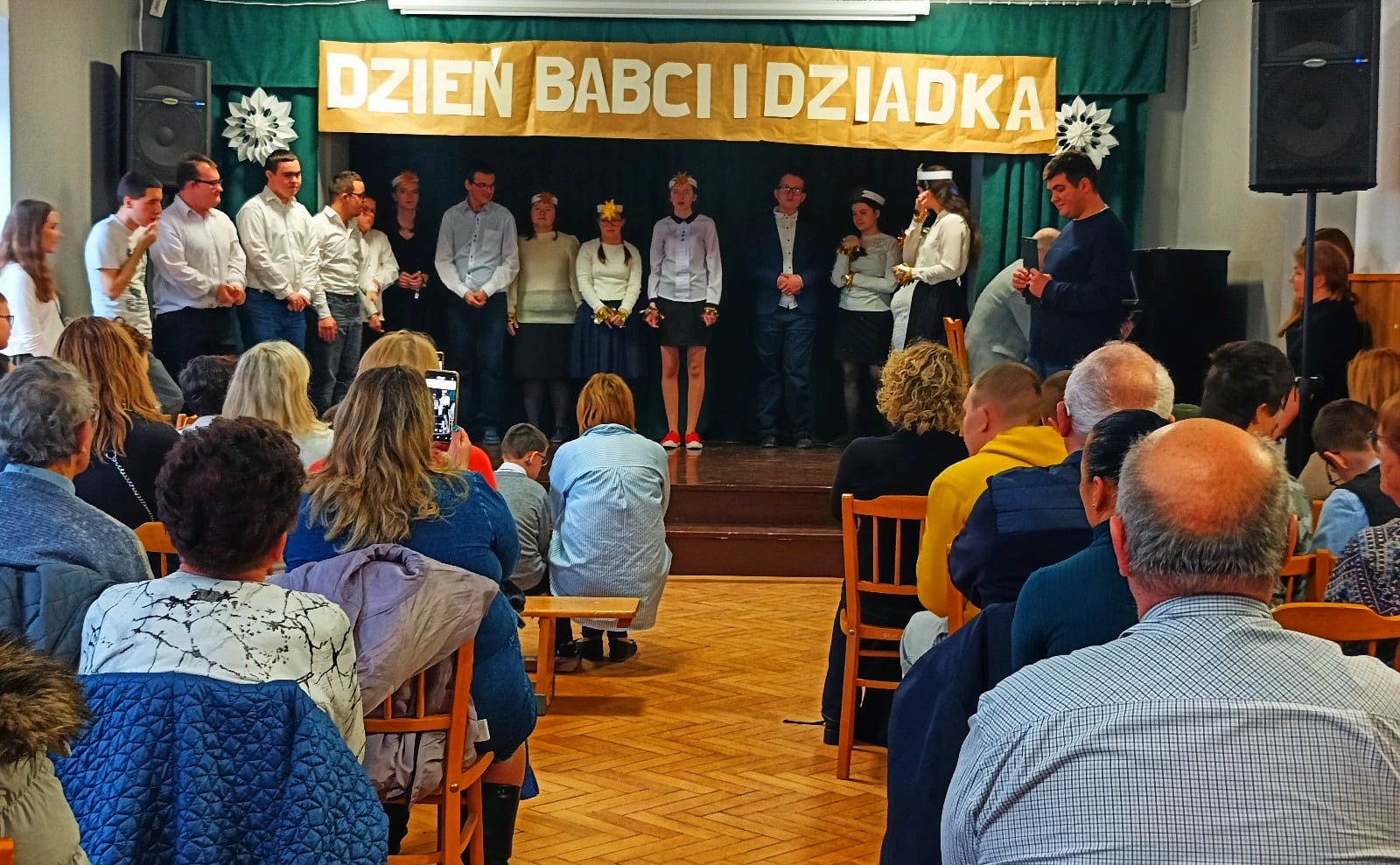 Uczniowie stoją na scenie w sali teatralnej, wystepują z okazji Dnia Babci i Dziadka.