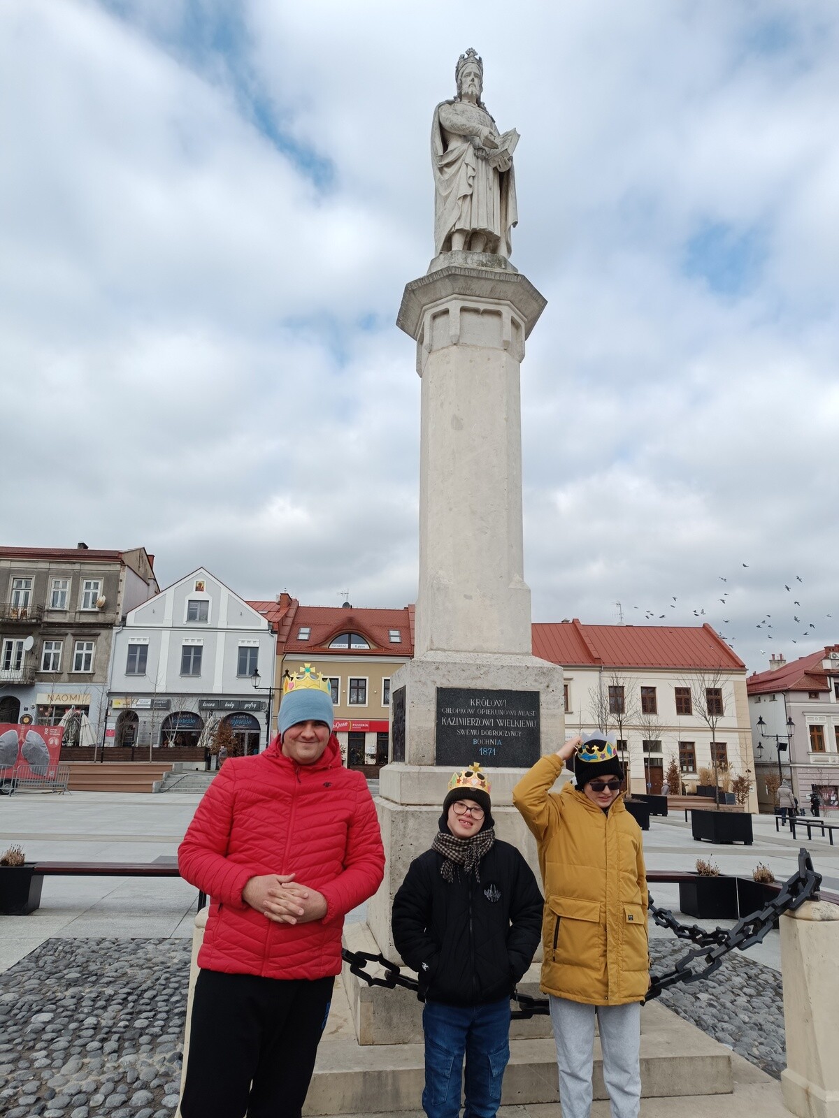 Trzech chłopców stoi na płycie rynku bocheńskiego, w tle widac pomnik Króla Kazimierza Wielkiego.