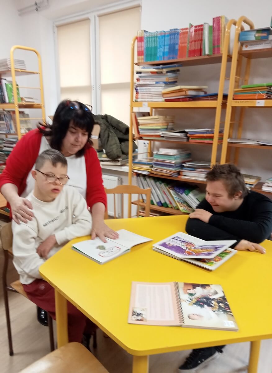 Dwoje uczniów siedzi przy stoliku w bibliotece szkolnej. Nauczyciel pochyla się nad stolikiem i pokazuje obrazek w książce, w tle widać regały z książkami.