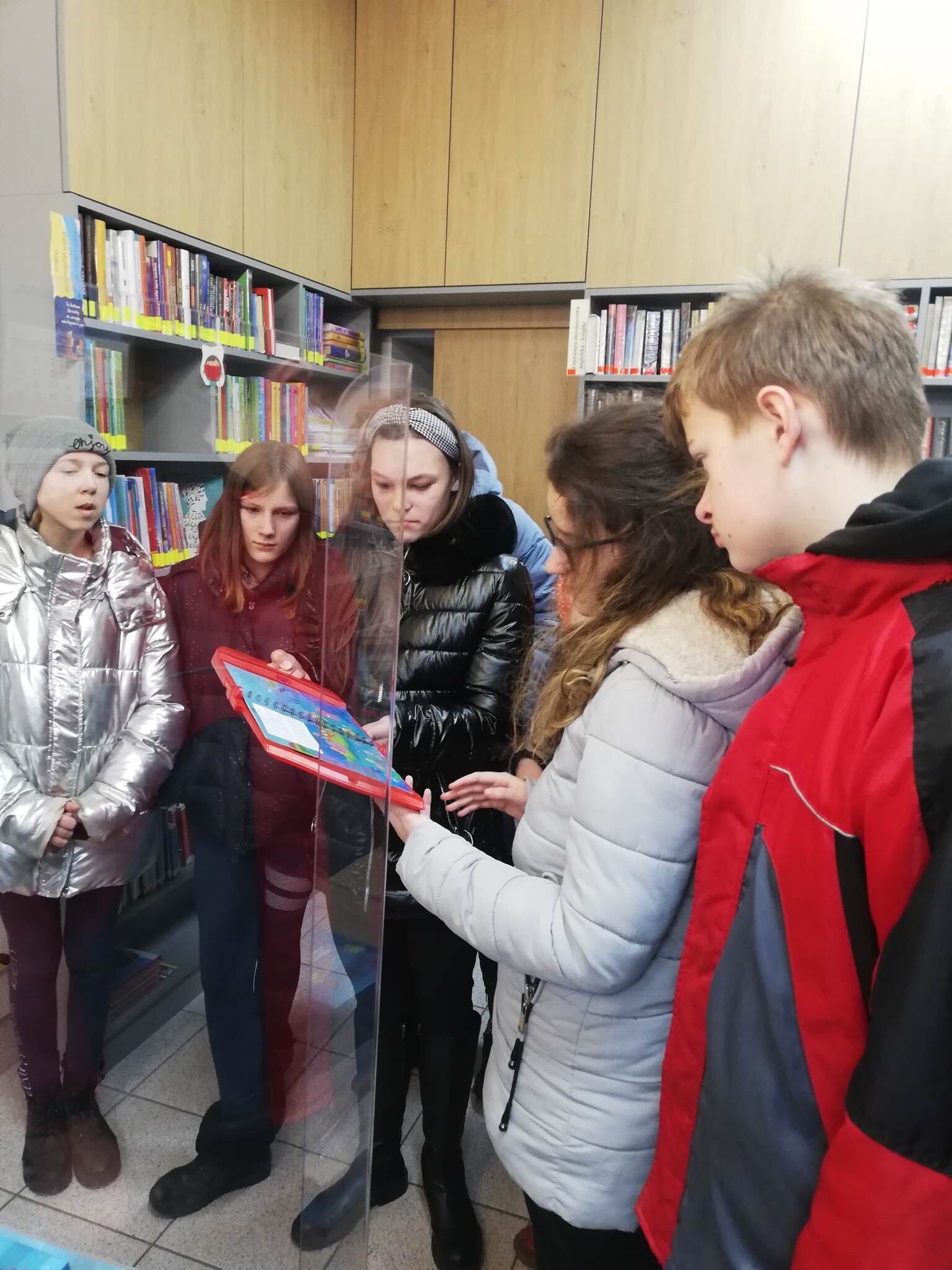 Uczniowie zwiedzają pomieszczenie biblioteki, spoglądają na regały z książkami.