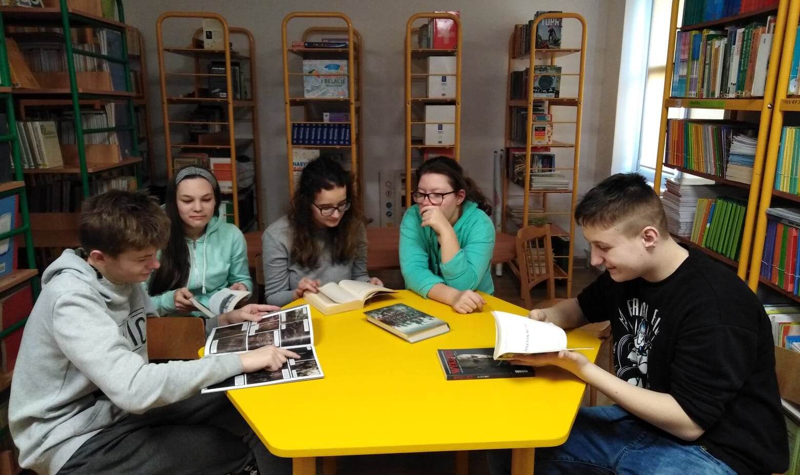 Uczniowie siedzą przy żółtym stoliku w bibliotece szkilnej, przeglądaja książki. W tle widać regały z książkami.