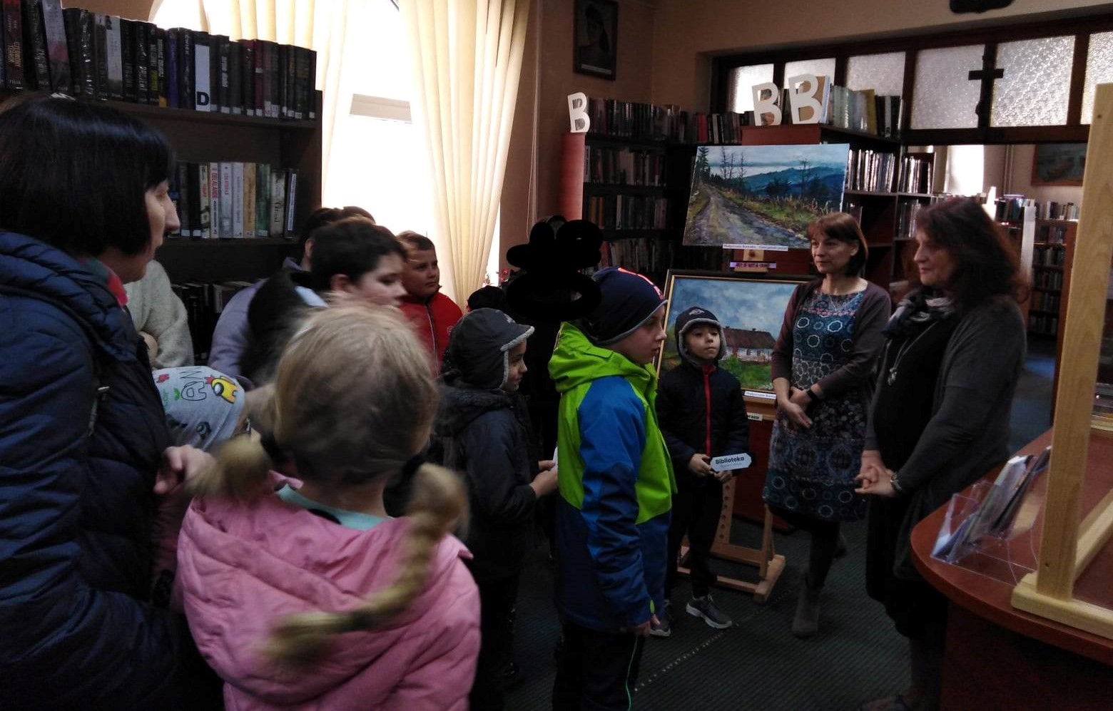 Uczniowie w pomieszczeniu biblioteki publicznej w Bochni stoją i słuchają wykładu kobiety pracującej w bibliotece.