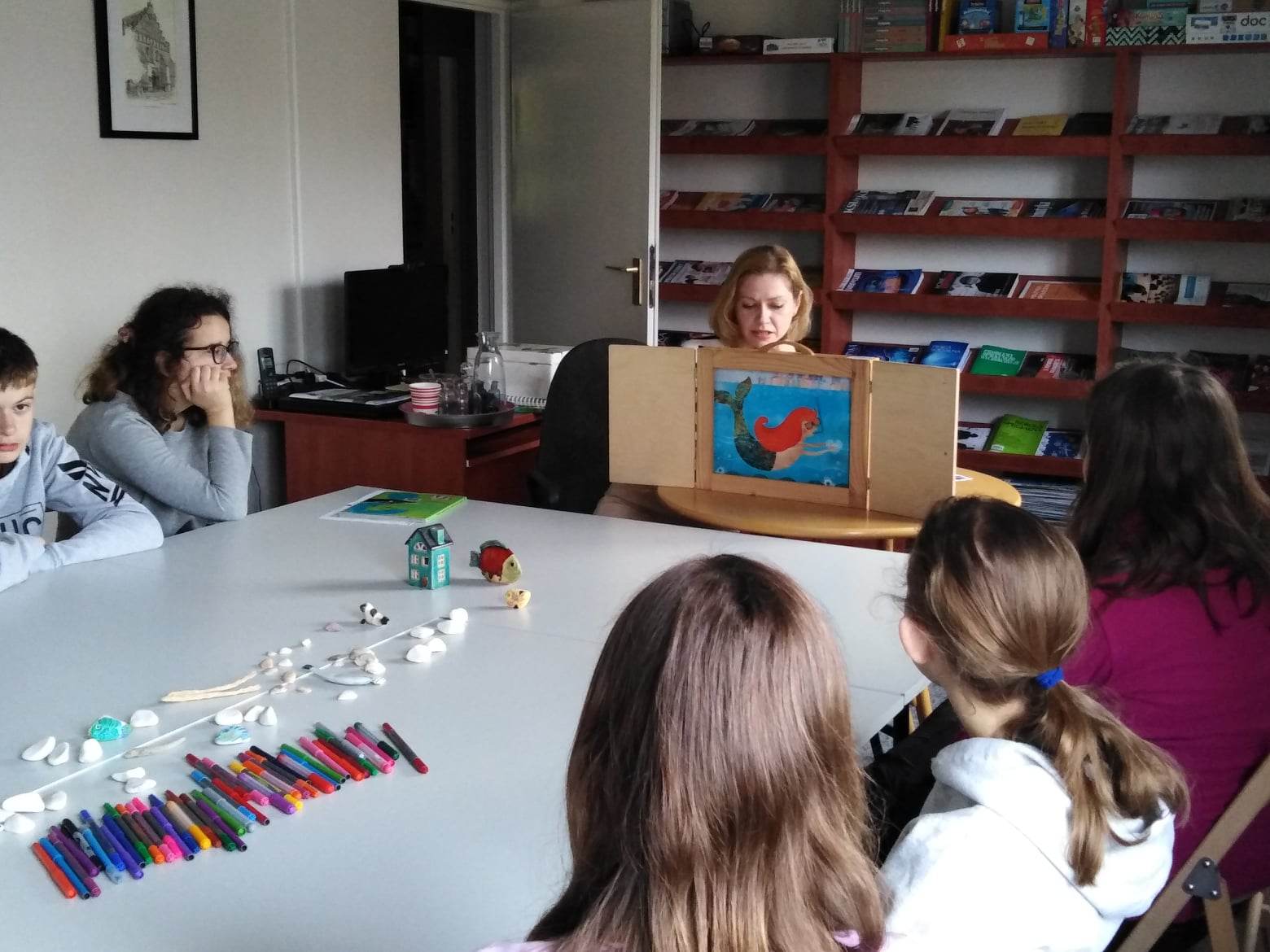 Uczniowie siedzą przy stole, patrzą w kierunku kobiety, która pokazuje ilustracje w drewnianej ramce.