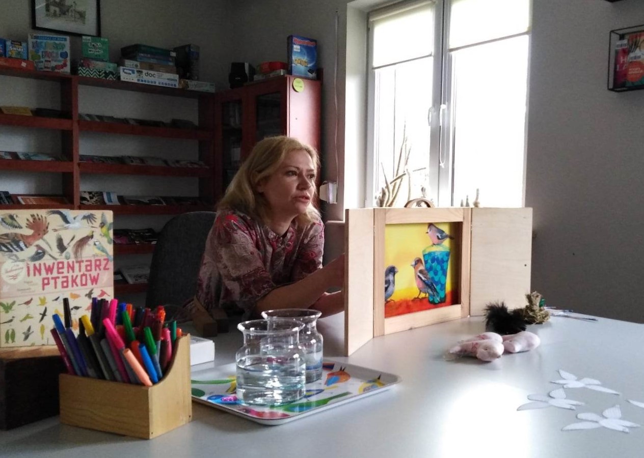 Kobieta w bibliotece siedzi przy stole, w dłoniach trzyma drewniana ramkę w której jest ilustracja dwóch ptaszków.