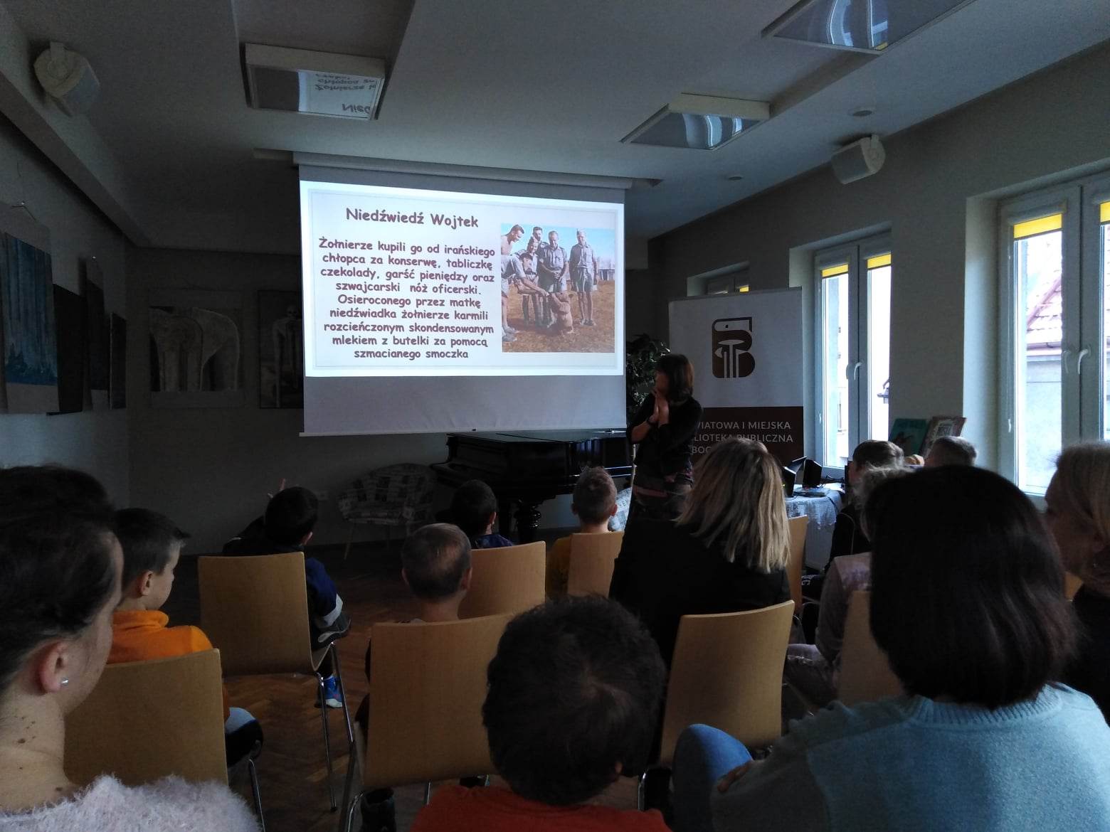 Grupa uczniów i nauczycieli siedzi na krzesłach, oglądają prezentację o Światowym Dniu Misia.