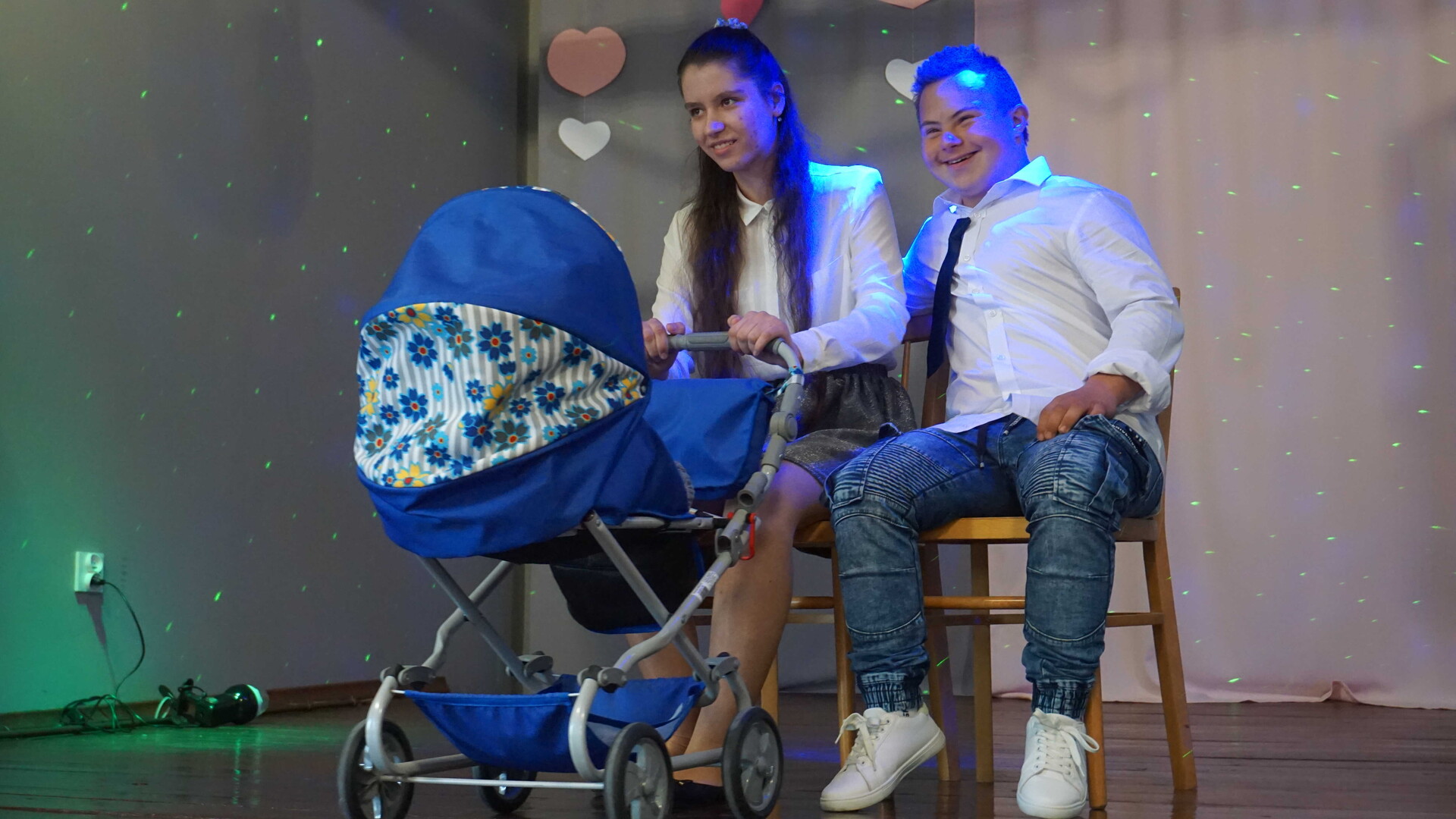 Chłopiec z dziewczyną siedzą na krzesłach na scenie, przed nimi stoi wózek dziecięcy. Oboje uśmiechają się.