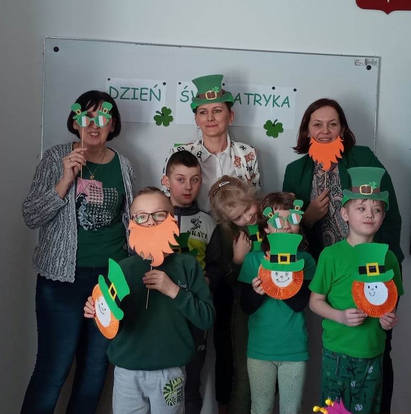 Uczniowie wraz z nauczycielem ubrani na zielono pozują do zdjęcia, w dłoniach trzymają wykonane z papieru. twarze skrzatów w zielonych kapeluszach.