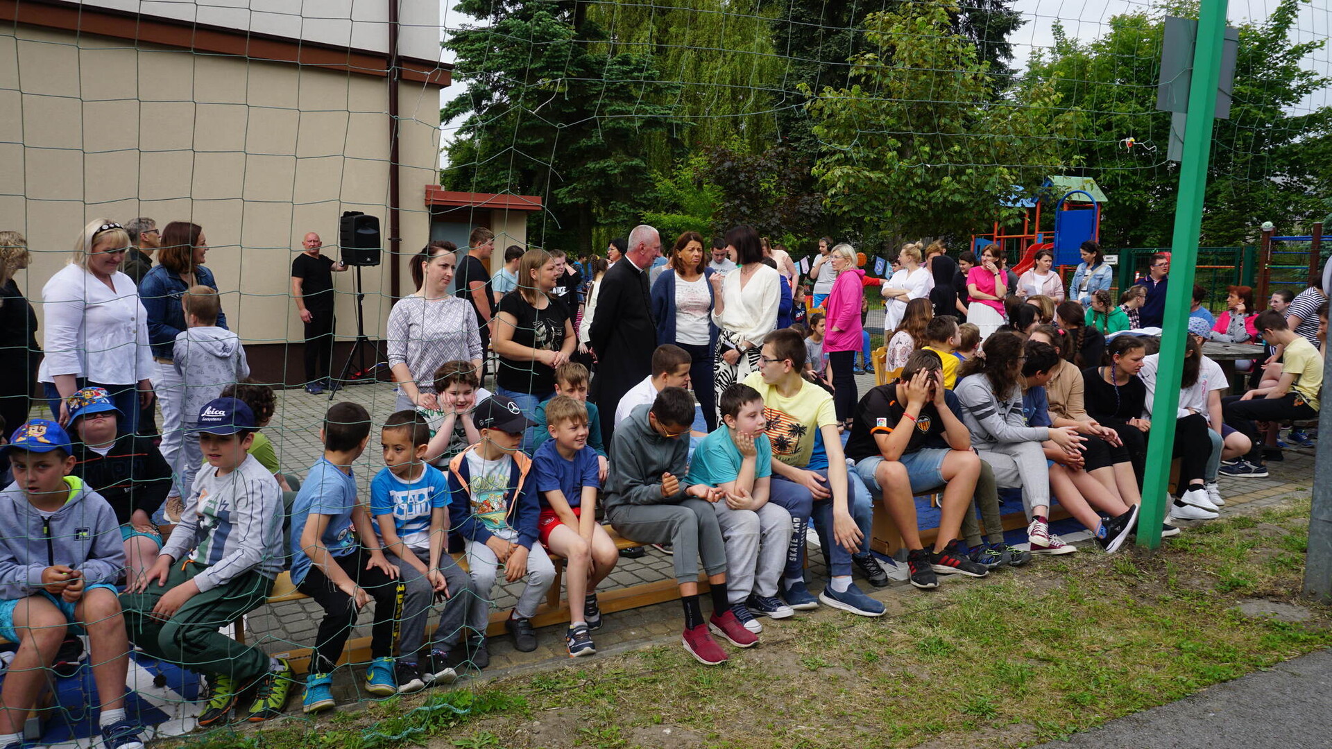 Na placu szkolnym, duża liczba uczniów w sportowych strojach siedzi na ławeczce, za nimi stoją kolejni uczniowie wraz z nauczycielami.