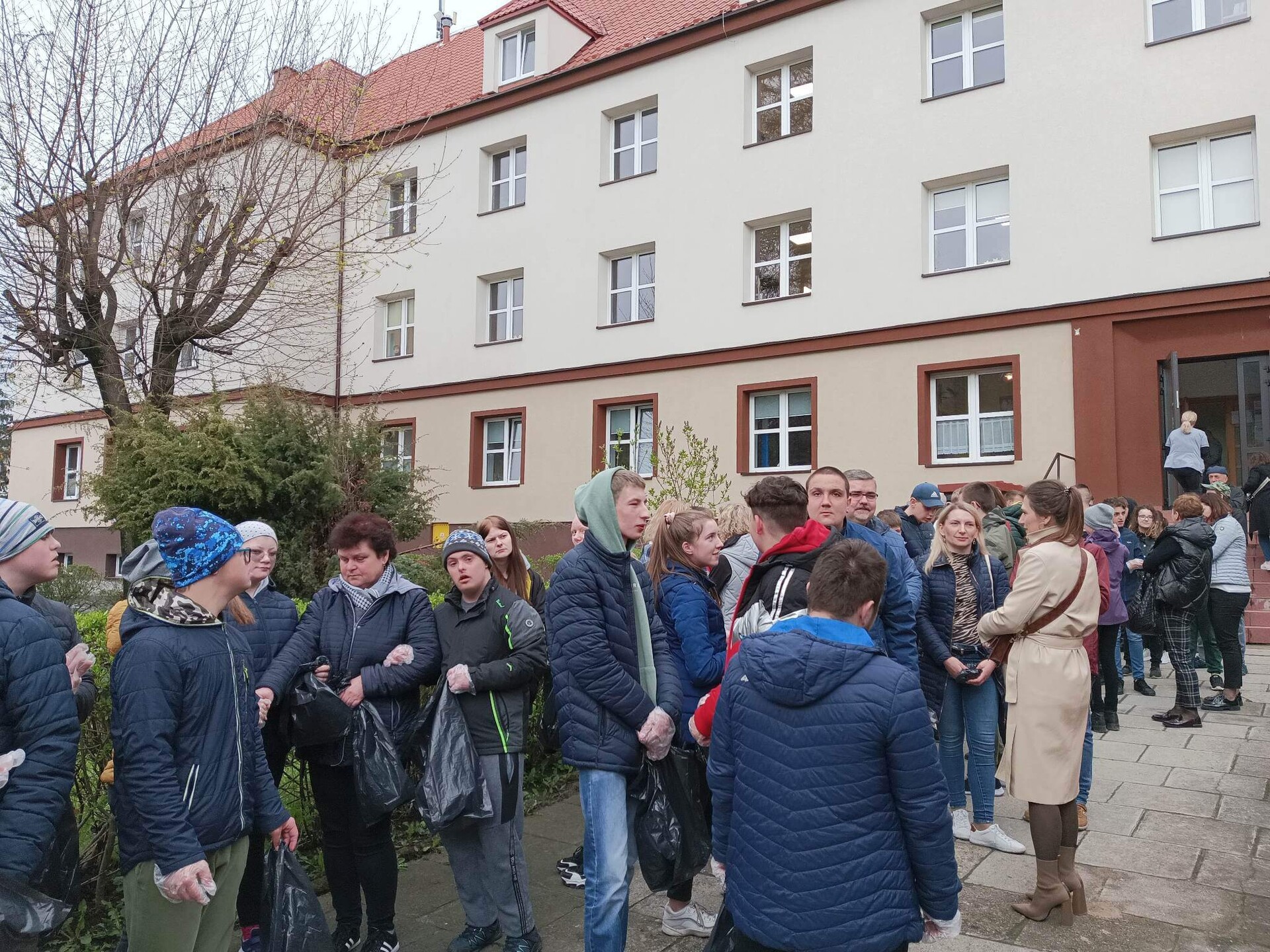 Grupa ludzi stoi na zewnątrz, obok budynku, w dłoniach trzymają czarne, foliowe worki na śmieci.