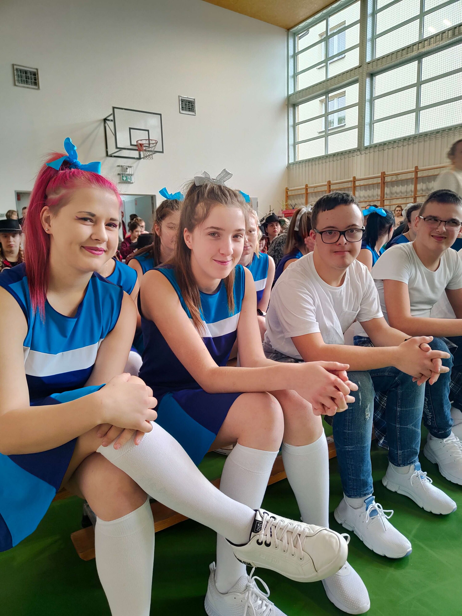 Uczniowie w niebieskich strojach tanecznych siedzą na ławeczce i pozują do zdjęcia w hali sportowej.