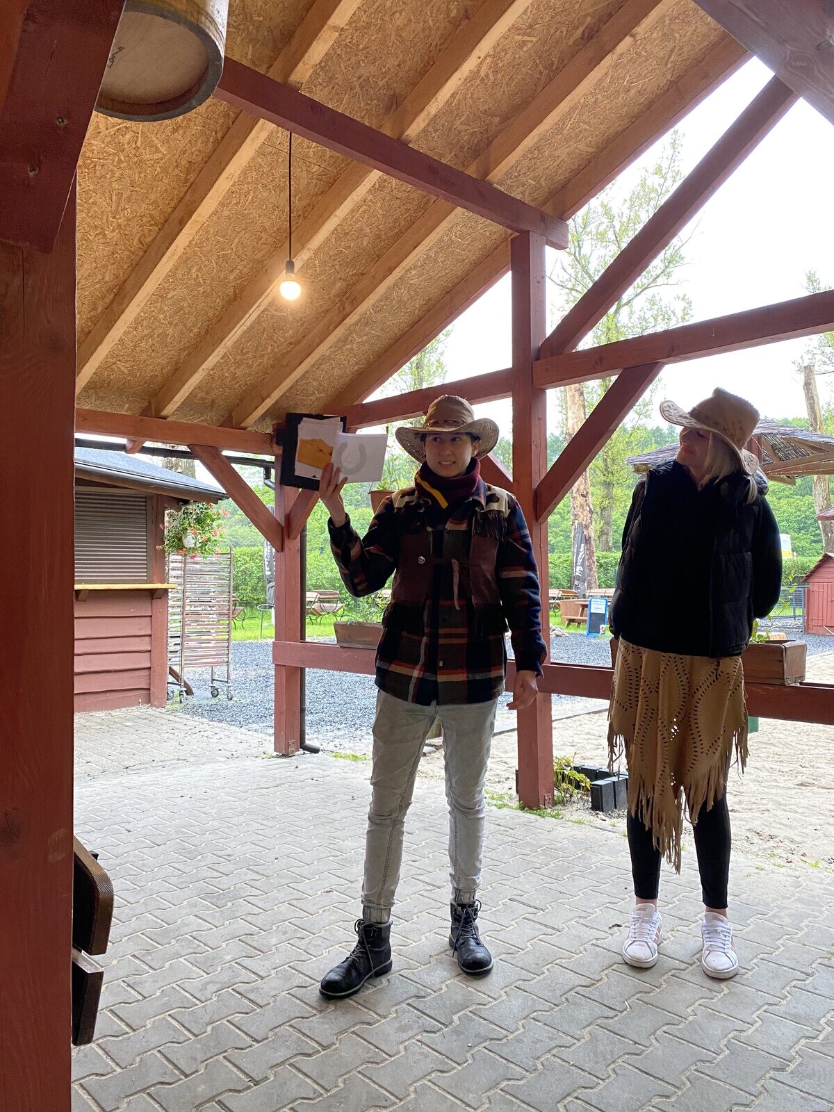 Dwie kobiety w kowbojskich kapeluszach i kurtkach z frędzlami stoją pod drewnianą  wiatą.