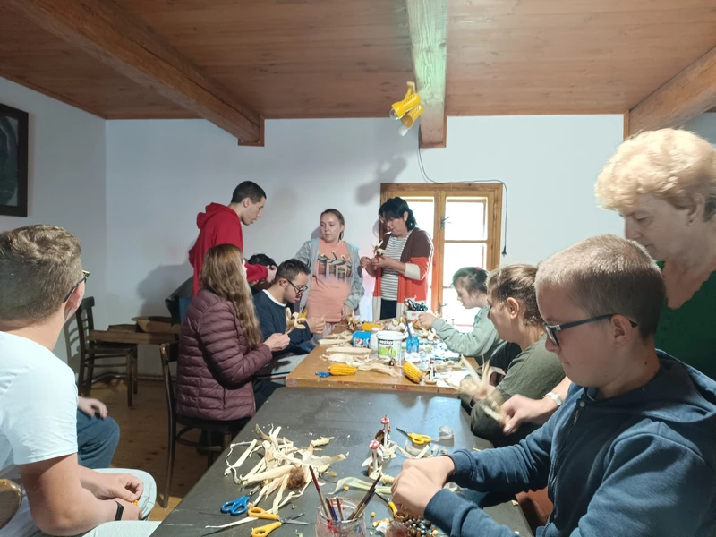 Grupa uczniów i nauczycieli znajduje sie w poieszczeniu. Częśc z nich siedzi rzy stole i wykonuje ozdoby z  kukurydzy. Pozostali  stoją i również wykonują rękodzieło z kukurydzy.