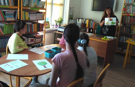 uczennice siedzą przy stoliku w bibliotece, patrzą w stronę bibliotekarza pokazującego ilustracje jabłka w książce.