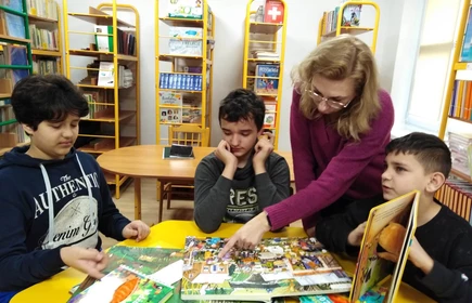 Troje uczniów siedzi przy żółtym stoliku w bibliotece szkolnej. Na stole leżą książki, w których pokazuje coś nauczyciel pochylający się nad uczniami.