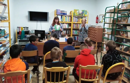 Uczniowie siedzą na krzesłach w bibliotece szkolnej, patrzą na kobietę trzymająca książkę. Obok stojąca kobieta wykonuje gest dłońmi, tłumaczy na język migowy.