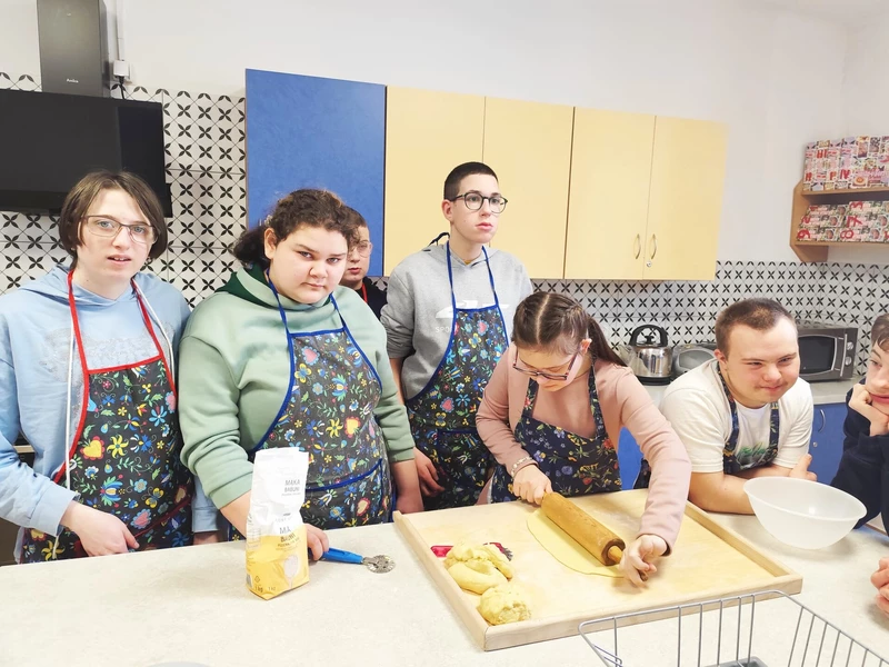 Uczniowie ubrani w fartuchy kuchenne stoją przy blacie, jedna dziewczynka wałkuje ciasto.