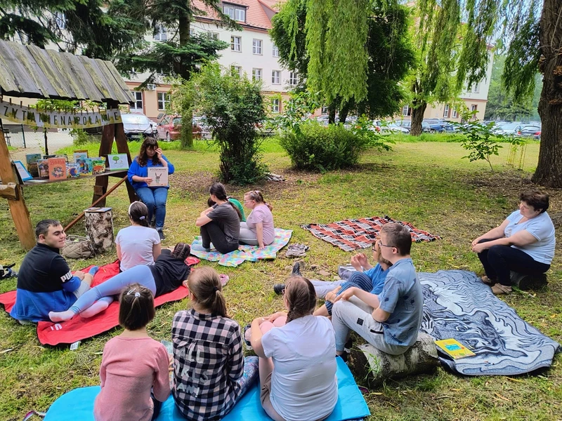 Grupa uczniów siedzi na trawie i słucha treści czytanej książki.