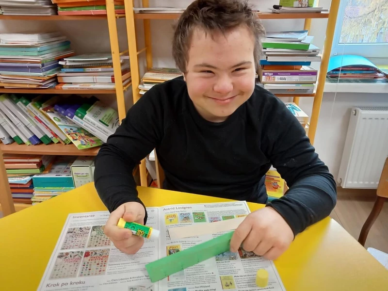 Chłopiec siedzi przy stoliku w bibliotece szkolnej, uśmiecha się, w te widać regały z książkami.