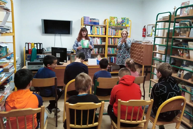 Uczniowie siedzą na krzesłach w bibliotece szkolnej, patrzą na kobietę trzymająca książkę. Obok stojąca kobieta wykonuje gest dłońmi, tłumaczy na język migowy.