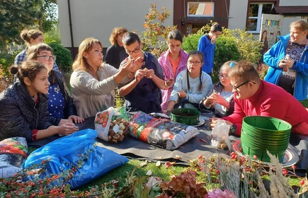 Grupa uczniów stoi wokół stołu w ogrodzie, patrza na nauczycielkę, która pokazuje nasiona.