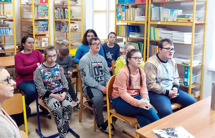 Grupa uczniów siedzi w bibliotece, w tle widać regały z książkami.