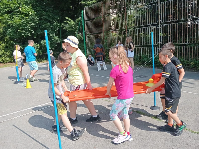 Czwórka dzieci niesie deskę ratowniczą, pokonują slalom między tyczkami na boisku szkolnym.