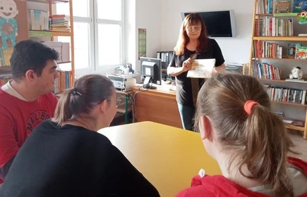 Uczniowie siedzą w bibliotece szkolnej i spoglądają w kierunku nauczyciela prezentującego książkę.