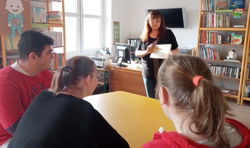 Uczniowie siedzą w bibliotece szkolnej i spoglądają w kierunku nauczyciela prezentującego książkę.