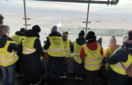 Grupa uczniów w odblaskowych kamizelkach odwrócona tyłem ogląda start samolotu z tarasu widokowego na lotnisku w Balicach.