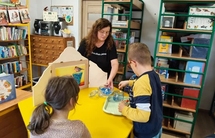 W bibliotece szkolnej nauczyciel pokazuje bajkę japońską dzieciom, które siedzą przy żółtym stoliku.