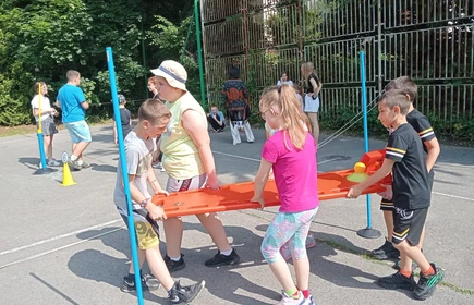 Czwórka dzieci niesie deskę ratowniczą, pokonują slalom między tyczkami na boisku szkolnym.