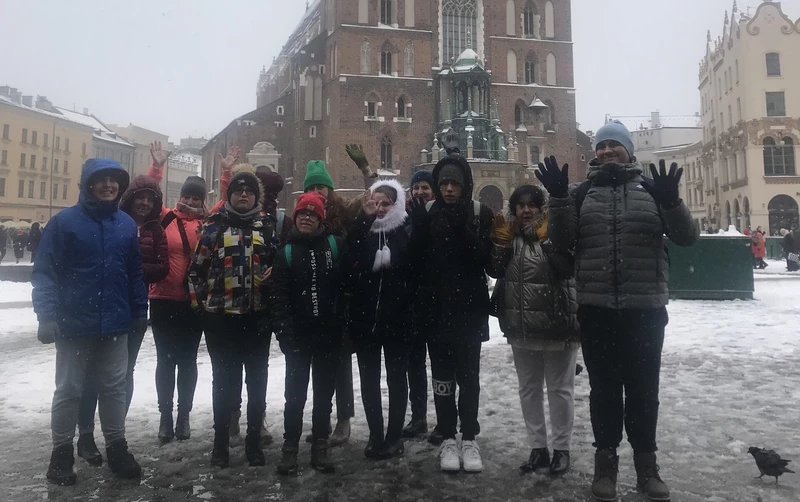 Grupa młodzieży ubranych w  grube czapki i kurtki pozuje do zdjęcia na tle Kościoła Mariackiego na RYnku Głównym w Krakowie, w zimowej scenerii.