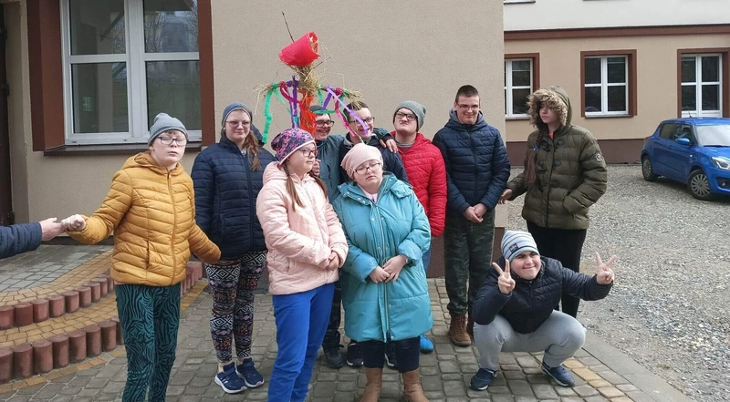 Uczniowie w ciepłej odzieży wierzchniej pozują do zdjęcia na tle budynku szkoły. Jeden uczeń trzyma w ręku kukłę Marzanny.