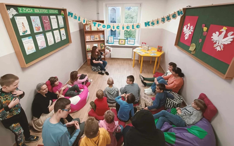 Grupa uczniów siedzi na podłodze, patrzą w kierunku kobiety, która czyta książkę i pokazuje obrazki w niej.