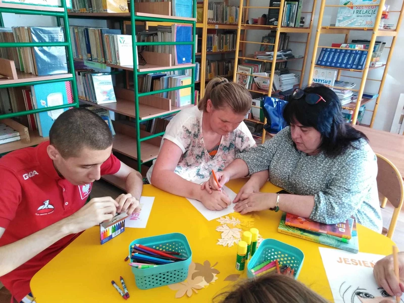 Uczniowie z nauczycielem siedzą przy stoliku w bibliotece szkolnej.  Na stoliku leżą ułożone książki.